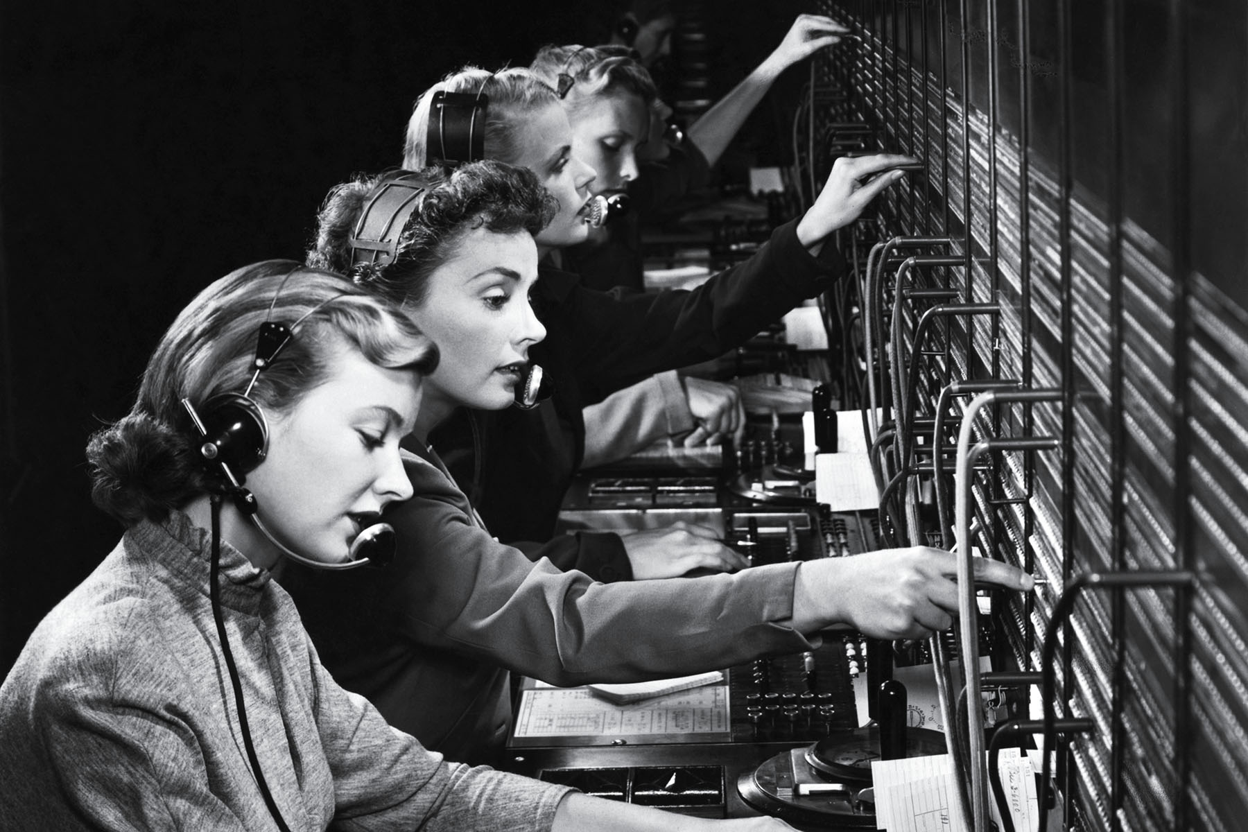 Women are seen from the side, operating a switchboard, in a black and white photo.