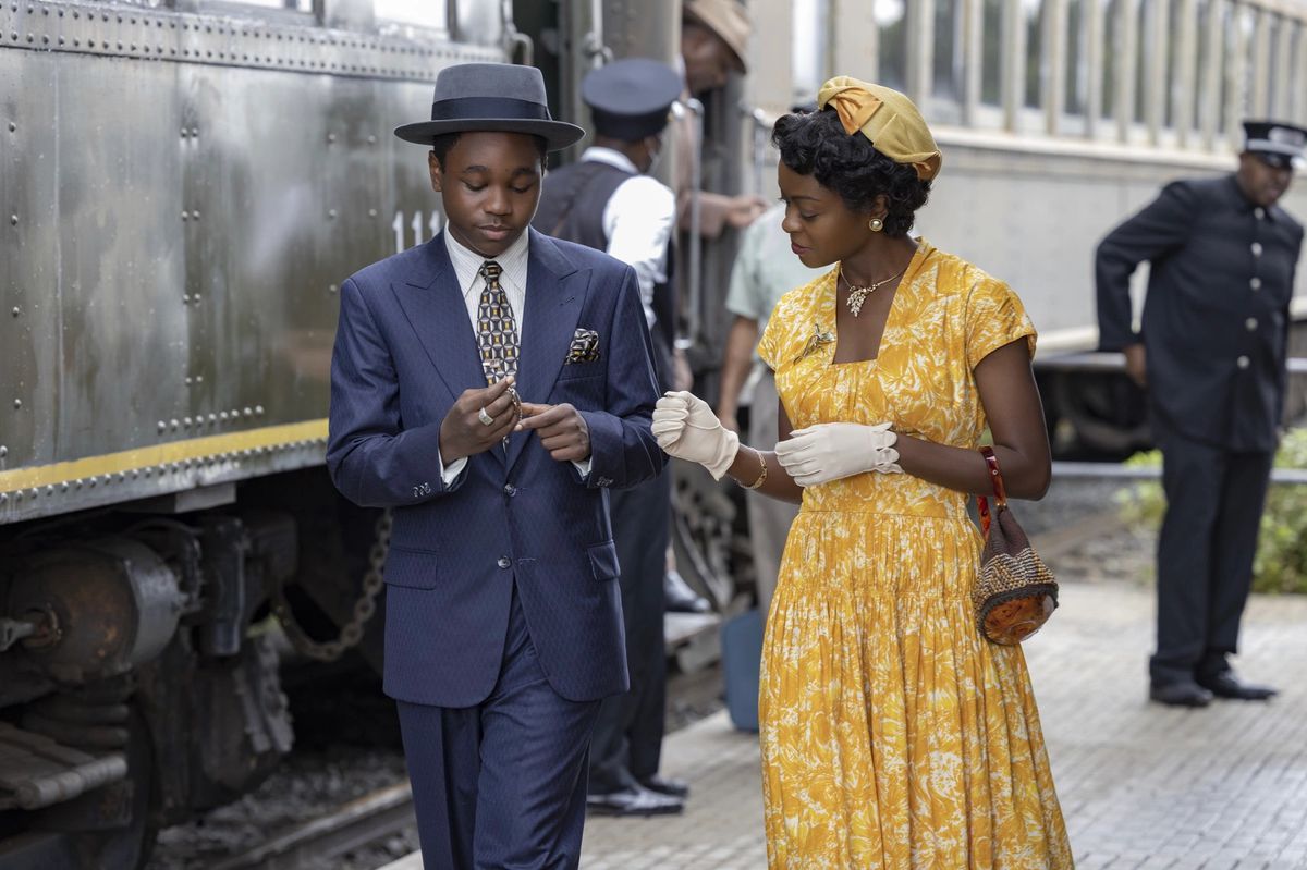 A boy in a navy blue suit (Jalyn Hall) stands next to a woman in a yellow dress (Danielle Deadwyler) beside a locomotive.