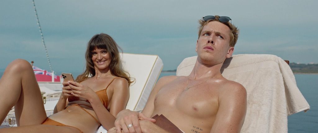 A smiling young woman in an orange bathing suit (Charlbi Dean) holds a cell phone and reclines on a white chair next to a bare-chested man (Harris Dickinson) in a bathing suit.