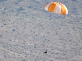 a small capsule descends toward a desert landscape beneath an orange and white parachute.